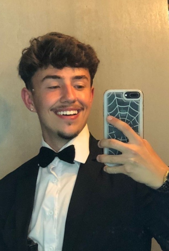 Jovem usando jaqueta de couro escura e camiseta, tirando selfie com smartphone em fundo escuro. Estilo casual e moderno.